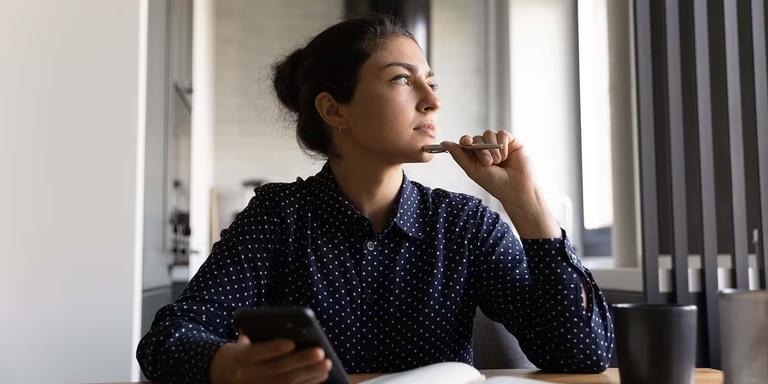 Professional woman sitting by a window, holding a pen and phone, looking thoughtful as she reflects on career decisions.