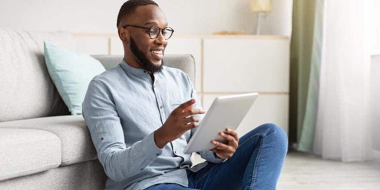 A smiling man sits on a floor leaning against a sofa, using a tablet to apply for jobs with AI tools.