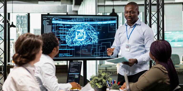 Team of tech professionals discuss AI development and related tasks during a meeting, with a screen showing an AI brain graphic and code.